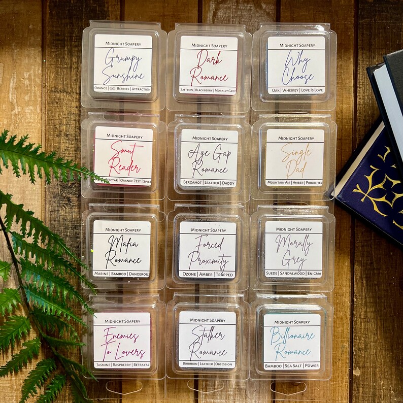 May include: Twelve small square containers of wax melts, each with a different scent and name. The names are all romance-themed, such as "Grumpy Sunshine", "Dark Romance", "Why Choose", "Smut Reader", "Age Gap Romance", "Single Dad", "Mafia Romance", "Forced Proximity", "Morally Grey", "Enemies to Lovers", "Stalker Romance", and "Billionaire Romance".