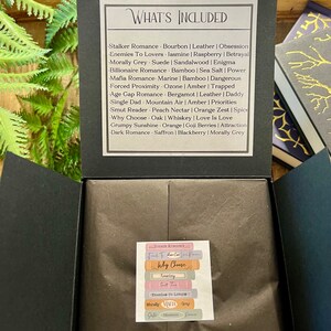 May include: A black box with a white label that says "What's Included" and lists different romance novel tropes with associated scents. The label also includes the text "Stalker Romance", "Enemies To Lovers", "Morally Grey", "Billionaire Romance", "Mafia Romance", "Forced Proximity", "Age Gap Romance", "Single Dad", "Smut Reader", "Why Choose", "Grumpy Sunshine", and "Dark Romance".