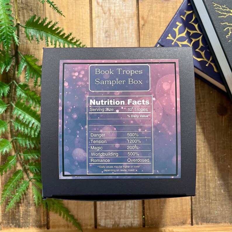 May include: A black box with a label that reads "Book Tropes Sampler Box" and a nutrition facts label listing tropes like "Danger", "Tension", "Magic", "Worldbuilding", and "Romance".