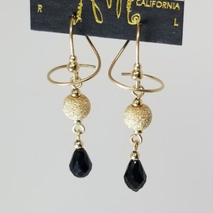May include: Gold tone earrings with a spiral design and a black teardrop-shaped bead. The earrings are on a black card with the text "HARRY MASON" and "CALIFORNIA" printed on it.