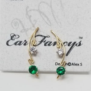 Op de afbeelding: Een paar gouden oorbellen met groene en witte edelstenen. De oorbellen zijn gevormd als een tak met een klein groen edelsteen aan het einde en een wit edelsteen in het midden. De oorbellen liggen op een witte kaart met de tekst "Ear Fancys" en "Harry Mason Presents".