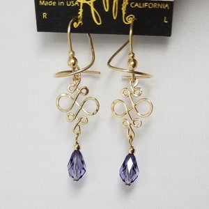 May include: Gold earrings with a delicate, twisted design and a dangling purple crystal. The earrings are on a black card with the text "HARRY MASON DESIGNER JEWELRY" and "CALIFORNIA".