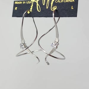 May include: A pair of silver hoop earrings with a twisted design and a small clear crystal in the center of each hoop. The earrings are hanging on a black card with the text "HARRY MASON" and "CALIFORNIA" printed on it.