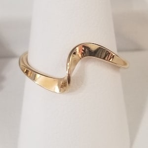 May include: A gold-toned ring with a wavy, abstract design. The ring is on a white display stand.