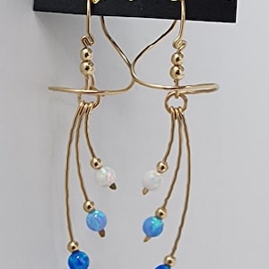 May include: Gold-toned dangle earrings with a delicate design. Each earring features a series of small gold beads and dangling strands adorned with white and blue opal-like beads. The earrings are from Harry Mason, Made in USA, California.
