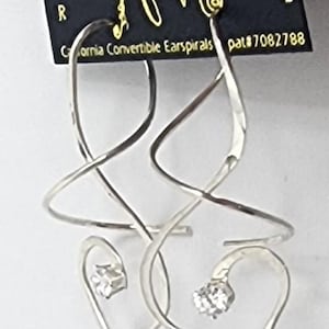 May include: Silver-toned convertible earspirals with a heart-shaped design, each featuring a small, sparkling stone. The earrings are from California and are marked with the text "Made in USA". The design includes the text "California Convertible Earspirals".