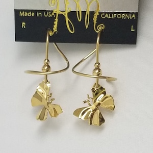 May include: A pair of gold-colored butterfly earrings. The earrings have a unique design with a spiral top and a butterfly pendant. The earrings are hanging on a black card that says "HARRY MASON" and "Made in USA".