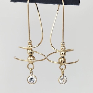 May include: Gold-tone earrings with a spiral design and a dangling, round, clear gemstone. The earrings have a post-back closure and are suspended from a curved wire. The earrings are on a black card with the text "HARRY MASON CALIFORNIA Made in USA".