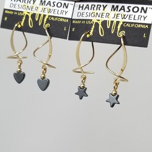May include: Two pairs of gold earrings with black gemstone pendants. One pair features a heart-shaped pendant and the other pair features a star-shaped pendant. The earrings are displayed on a black card with the text "HARRY MASON™ DESIGNER JEWELRY" and "CALIFORNIA".