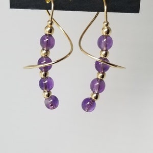 May include: A pair of gold earrings with a dangling purple amethyst stone. The earrings are made of wire that is twisted into a spiral shape. The earrings are labeled "Harry Mason" and "California".