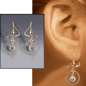 May include: Gold earrings with a dangling circle and a small clear stone. The earrings are shown on a model's ear.