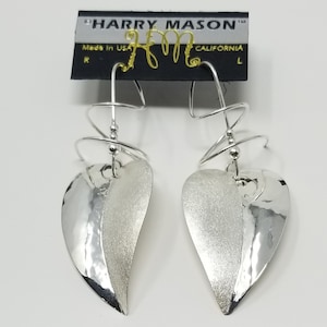 May include: Silver heart-shaped earrings with a textured finish. The earrings hang from a silver wire that is twisted into a spiral shape. The earrings are labeled "Harry Mason" and "Made in USA, California".