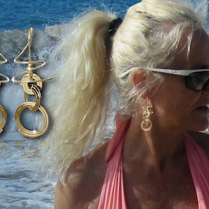 May include: Gold earrings with a geometric design. The earrings feature multiple interlocking circles and a curved wire design. The earrings are shown against a background of blue water and white waves.