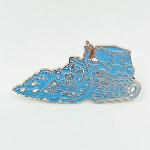 May include: A blue and gold enamel pin of a bulldozer with a wave of water behind it. The bulldozer has a small figure of a person in the cab.
