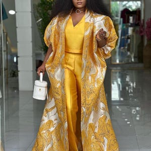 Puede incluir: Una mujer con un traje de pantalón amarillo y una chaqueta larga con estampado dorado y blanco. Lleva gafas de sol y un collar de oro.