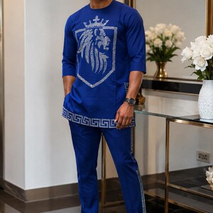 May include: A royal blue two-piece outfit consisting of a round-neck tunic and matching trousers. The tunic features a large silver rhinestone lion head emblem on the chest and a geometric Greek key pattern border in silver rhinestones at the hem.