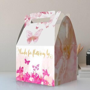 May include: A white favor box with a pink and gold butterfly design. The box has a handle and says "Thanks for fluttering by".