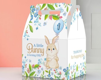 May include: A white favor box with a blue and green floral design. The box features a cartoon bunny holding a blue balloon with the number "1" on it. The text "A little Bunny is turning ONE!" is printed on the front of the box. The back of the box says "Thank you for hopping by".