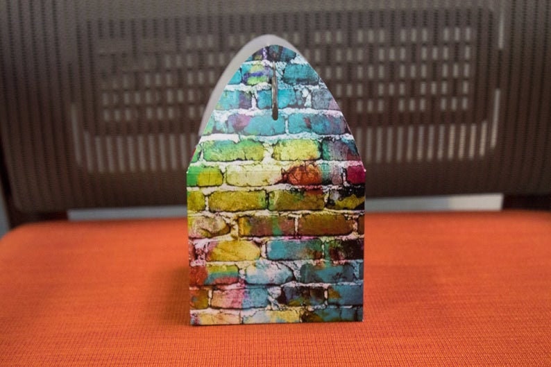 Puede incluir: Una peque&ntilde;a caja colorida con un dise&ntilde;o de patr&oacute;n de ladrillo. La caja est&aacute; hecha de cart&oacute;n y tiene una solapa en la parte superior.