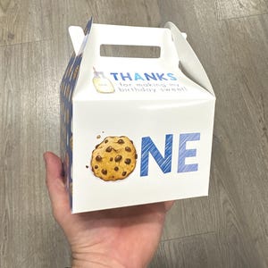 May include: A white favor box with a blue and white cookie pattern. The box has a handle and says "THANKS for making my birthday sweet! ONE" with a drawing of a chocolate chip cookie.