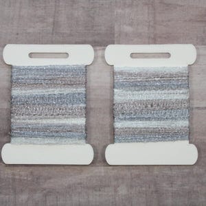 May include: Two spools of yarn with a white cardboard backing. The yarn is a mix of blue, silver, and brown, with a textured appearance. The spools are side-by-side against a neutral background.