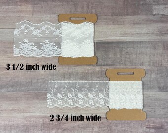 May include: Two spools of white floral lace trim. The top spool features a scalloped edge lace measuring 3 1/2 inches wide. The bottom spool features a straight-edged lace with a delicate vine pattern measuring 2 3/4 inches wide.