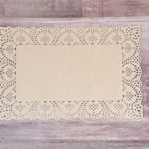May include: A rectangular, cream-colored paper doily with a decorative, openwork lace border. The doily is placed on a light brown wooden surface, showcasing its delicate design and vintage aesthetic. The doily's dimensions are approximately 8 x 10 inches.