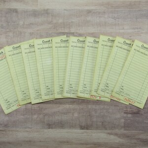 May include: A stack of pale yellow guest check pads with printed lines and the words "Guest Check" at the top. Each pad has a unique number printed in red. The pads are arranged on a light brown wooden surface.