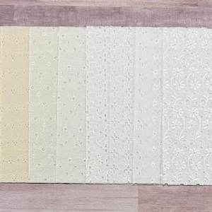 May include: A collection of 15 white fabric strips with different embroidered patterns. The fabric is a light cream color and the embroidery is in white thread.