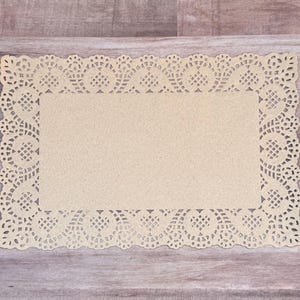 May include: A rectangular beige paper doily featuring a solid center and an intricate, die-cut lace border with repeating scalloped edges and geometric patterns. The paper has a textured, matte finish and a light tan color throughout the design.