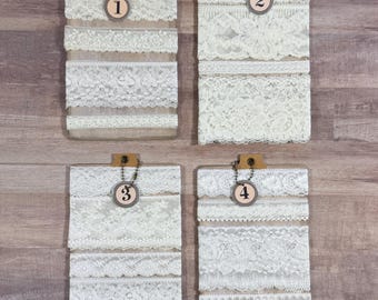 May include: Four clipboards displaying various white lace trims. Each clipboard has a small tag with a number from one to four. The lace trims vary in pattern and texture, showcasing a range of delicate designs. The clipboards are arranged on a light brown wooden surface.