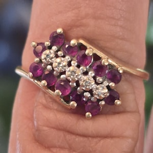 May include: A gold ring featuring a cluster of round, deep pink gemstones and smaller, clear, round stones arranged in a central row. The stones are set in a bypass-style gold band that curves around the cluster of stones on both sides.