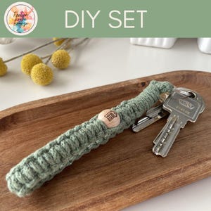 Könnte beinhalten: Ein DIY-Set mit einem salbeigrünen Makramee-Schlüsselanhänger mit Holzperle und Hausdesign. Der Schlüsselanhänger hält mehrere Schlüssel. Über dem Schlüsselanhänger steht der Text "DIY SET".