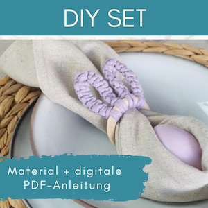 Könnte beinhalten: DIY-Set zum Erstellen von Serviettenringen. Das Set enthält Materialien und eine digitale PDF-Anleitung. Das Bild zeigt eine hellgraue Leinen-Serviette mit einem geflochtenen Ring in Lavendelfarbe, der darum gebunden ist. Die Serviette liegt auf einem weißen Teller mit einem geflochtenen Stroh-Untersetzer darunter.