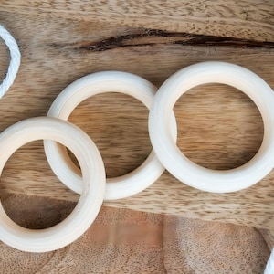 Könnte beinhalten: Draufsicht auf Holzturnringe und weißes Seil auf einer Holzoberfläche. Die Ringe sind hell und kreisförmig, mit glatter Oberfläche. Das Seil ist weiß und gedreht, was der Komposition Textur verleiht. Thema: Sport und Fitness.