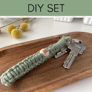 Könnte beinhalten: Ein DIY-Set mit einem salbeigrünen Makramee-Schlüsselanhänger mit Holzakzent und einem Schlüsselsatz, der auf einem Holztablett liegt. Der Text "DIY SET" befindet sich oben im Bild.