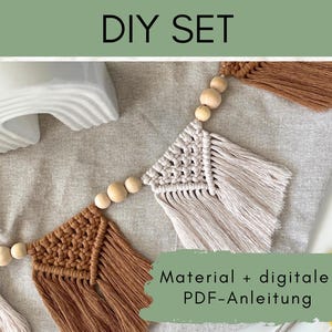Könnte beinhalten: Ein DIY-Makramee-Girlanden-Set mit Holzperlen und Fransen in Beige und Braun. Das Bild enthält den Text "DIY SET" und "Material + digitale PDF-Anleitung". Die Girlande wird auf einer neutralen Stoffoberfläche präsentiert.
