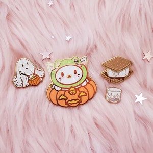 May include: Four enamel pins featuring Halloween-themed designs. A white ghost holding a pumpkin, a green frog wearing a pumpkin costume, a brown and white s'more, and a white marshmallow.