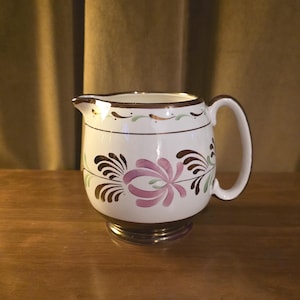 May include: A white ceramic pitcher with a brown rim and handle, featuring floral designs in pink, brown, and green. The pitcher has a decorative base and is likely used for serving beverages.