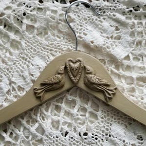 May include: A beige wooden clothes hanger with a metal hook. The hanger features a decorative design of two birds facing each other, with a heart in the center. The birds and heart are raised from the surface of the hanger.