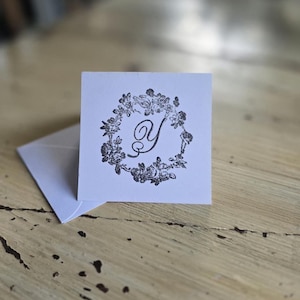 May include: A square white card featuring a black ink stamp of the letter Y centred within a detailed floral wreath design, accompanied by a matching white envelope. The card and envelope are made of paper with a clean, matte finish.