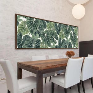 May include: A rectangular wall art piece featuring a dense, overlapping pattern of large, heart-shaped green leaves with prominent white veins. The artwork is framed in a thin, dark wood border and is displayed horizontally on a textured white wall.