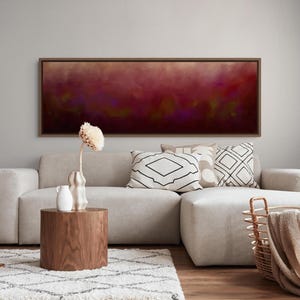 May include: A panoramic rectangular wall art piece featuring an abstract blend of deep burgundy, dark purple, and muted gold tones, enclosed in a thin brown frame. The artwork hangs above a light gray sectional sofa with patterned throw pillows.
