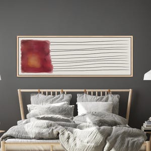 May include: Rectangular framed artwork with a cream background. A large, blurred burgundy and orange square is on the left, with horizontal black lines on the right. The artwork is displayed above a bed with gray bedding.