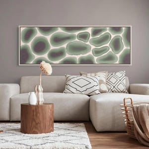 May include: A rectangular wall art piece featuring a repeating pattern of organic, rounded shapes in shades of sage green and deep purple against a cream background, enclosed in a thin white frame and displayed horizontally above a sofa.