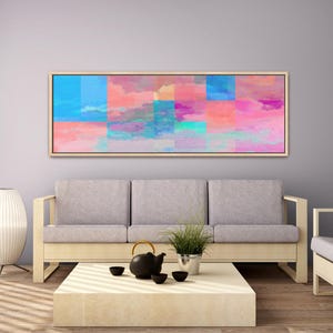 May include: A rectangular framed artwork with a colorful abstract design hangs above a gray sofa. The artwork features a patchwork of blues, pinks, and purples, resembling a cloudy sky. A light-colored wooden coffee table holds a black teapot and cups.