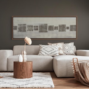 May include: A rectangular wall art piece featuring a series of gray square shapes on a light gray background, framed in a thin brown border. The artwork is displayed above a light beige sectional sofa with patterned throw pillows and a wooden side table.