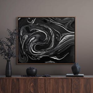 May include: Abstract black and white art print with swirling patterns. The artwork is framed in a wooden frame and displayed on a wooden cabinet. The color palette includes shades of black, white, and gray.