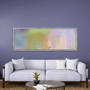 May include: Abstract art piece in a silver frame. The artwork features a blend of pastel colors, including blue, green, yellow, and pink. The painting hangs above a white sofa with pillows, a round coffee table, and a small plant.