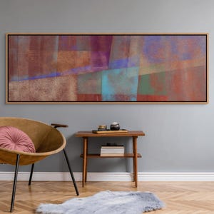 May include: Abstract art print in a wooden frame. The artwork features a blend of colors, including shades of red, purple, blue, and green. The rectangular piece is mounted on a gray wall above a mid-century modern style chair and side table.